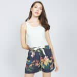 A person wearing a light-colored tank top and navy shorts with a tropical floral pattern. They are standing in front of a plain light gray background.